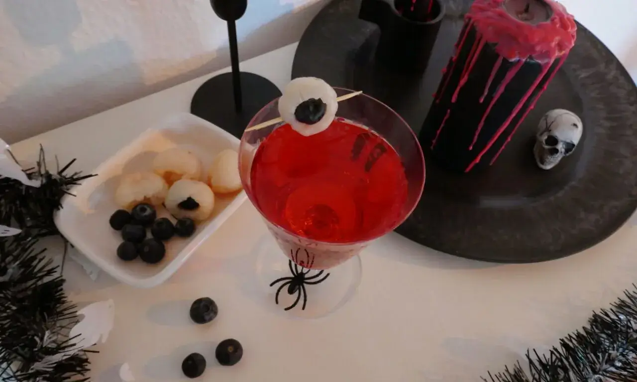 Halloween-Cocktail mit Litschi-Auge, roten Kerzen und Totenschädel-Deko Roter Halloween-Drink im Glas mit Spinnenmotiv, Litschi-Blaubeer-Auge als Deko, daneben Kerze mit Wachsspuren und Totenschädel.