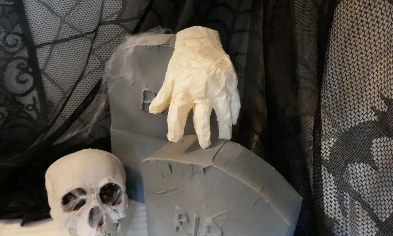 Halloween-Deko mit Grabsteinen, Totenschädel und ausgestreckter Geisterhand Halloween-Szene mit grauen Grabsteinen, weißem Totenschädel und einer gruseligen, ausgestreckten Hand auf dunklem Netz-Hintergrund.