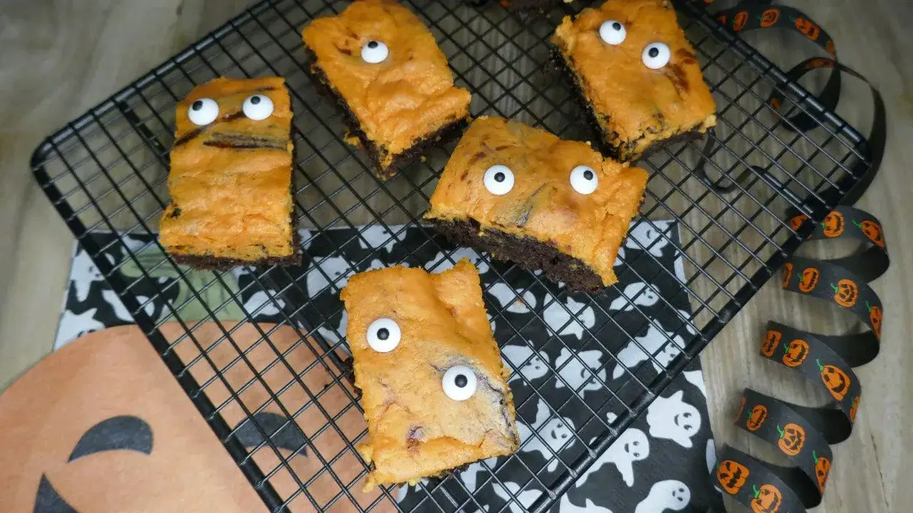 Halloween-Brownies mit Zuckerdekor-Augen auf schwarzem Kuchengitter Halloween-Brownies mit orangefarbener Oberfläche und Zuckerdekor-Augen auf Gitterrost, dekoriert mit Geisterserviette und Kürbisband.
