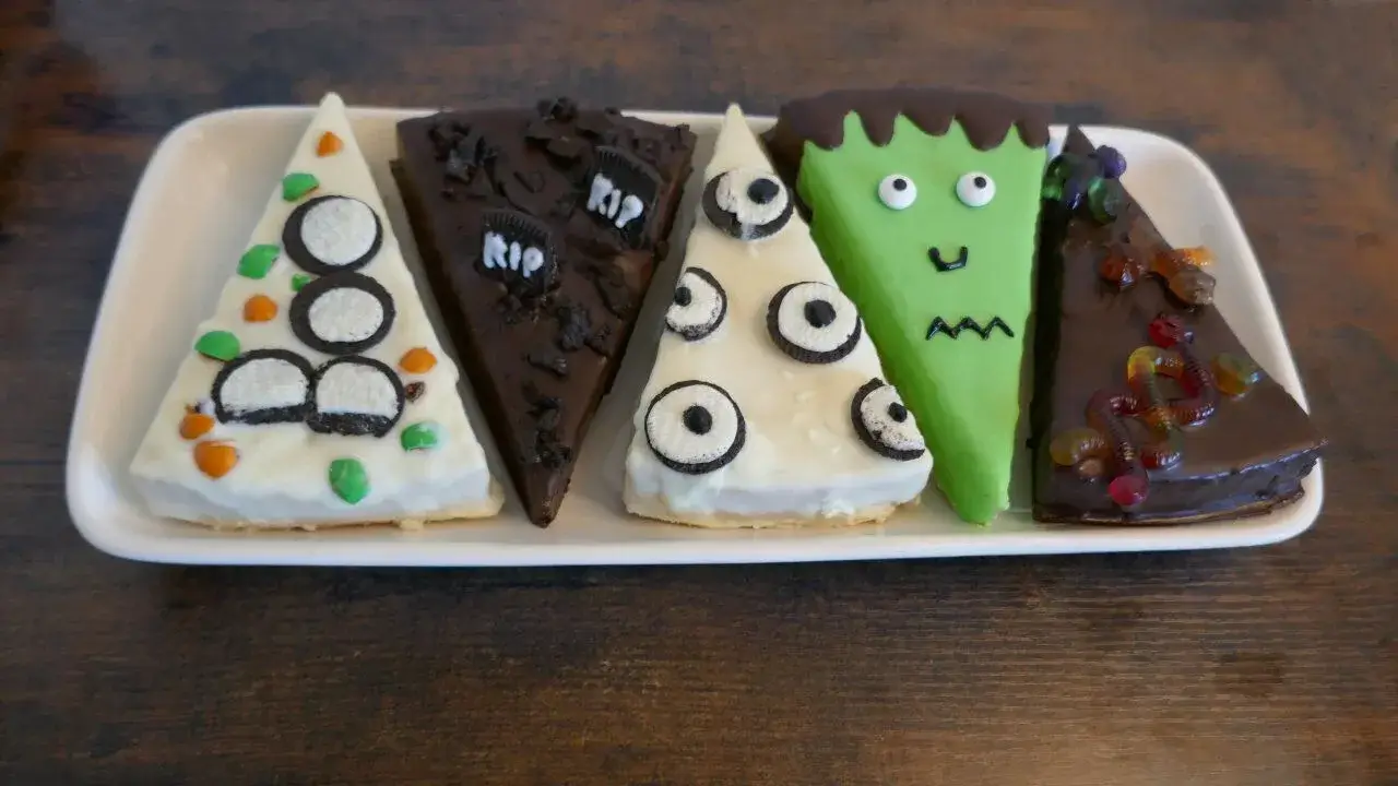 Halloween-Kuchenstücke mit kreativer Dekoration aus Oreo-Augen, Fondant und Gummiwürmern Fünf Kuchenstücke mit Halloween-Dekor: Geister, Augen, Frankenstein, Gummiwürmer und "RIP"-Gräber auf bunter Glasur.