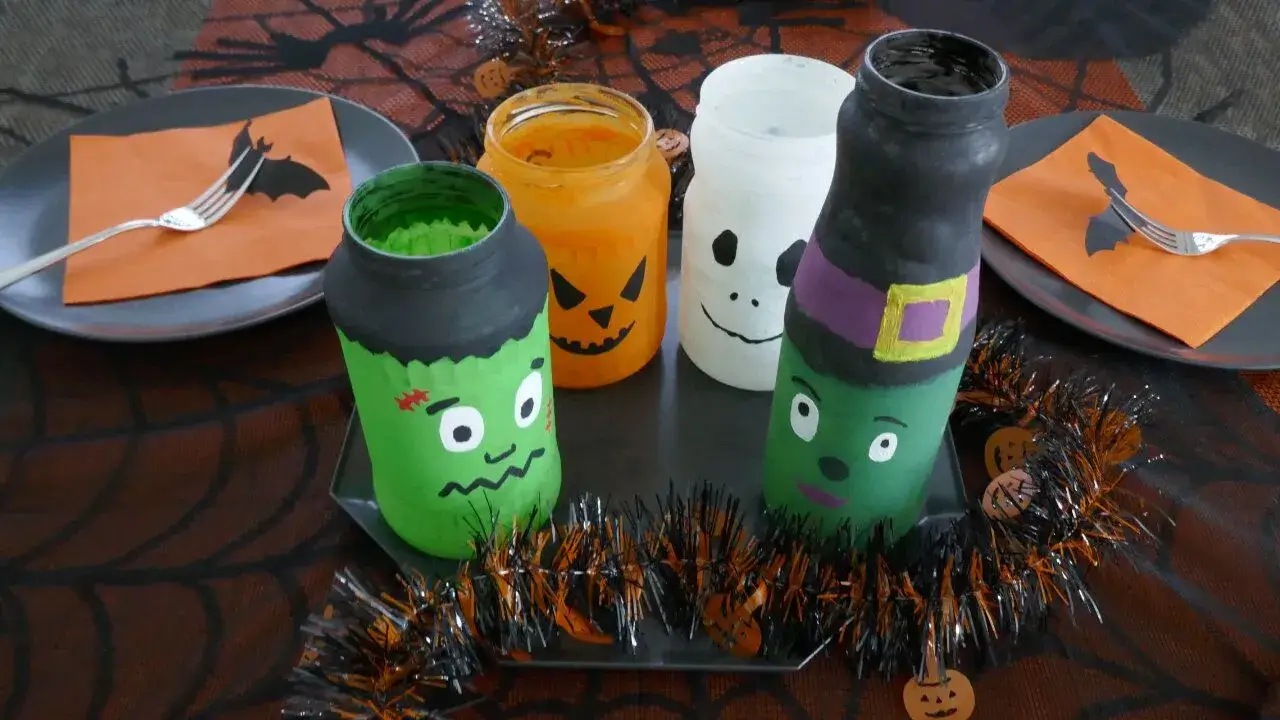 Halloween-Windlichter aus Gläsern mit bemalten Gesichtern von Kürbis, Geist, Hexe und Monster Vier bemalte Halloween-Windlichter aus Gläsern mit Kürbis-, Geist-, Frankenstein- und Hexengesicht auf festlich gedecktem Tisch.